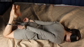 Anne- barefoot blindfolded Anne in grey dress, hogcuffed with steel handcuffs and legcuffs, rolls on the bed (HD WMV).webp