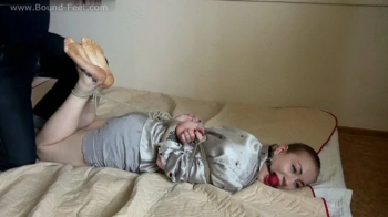 Ballgagged barefoot Amanda in grey shirt and shorts, hogtied with her thumbs tied up, is getting her feet oiled and tick.webp