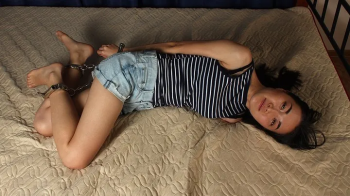 Carrie- barefoot girl in tank top and jeans shorts, hogcuffed with shiny steel cuffs, rolls and wriggles on the bed (HD.webp